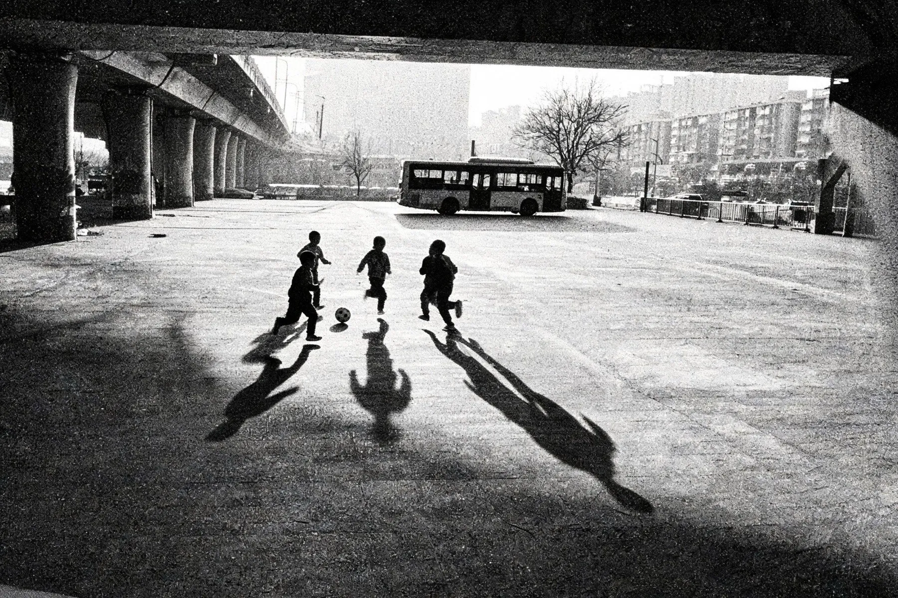 Vier kinderen rennen achter een bal aan onder een viaduct, met lange schaduwen op het beton